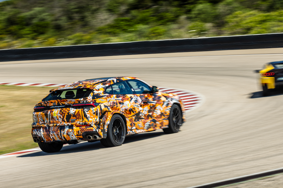 A camouflaged 2026 Lamborghini Urus Performante prototype seen from the rear, driving on a racing circuit with a yellow Lamborghini following behind.