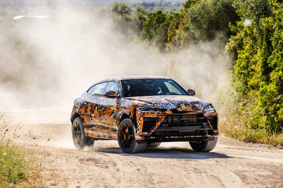 A Lamborghini Urus SUV prototype wrapped in orange camouflage livery driving on a dusty dirt road, kicking up a cloud of dust
