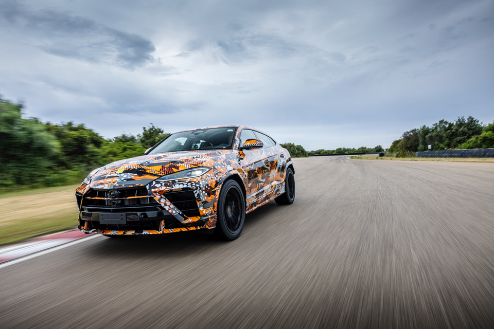 A camouflaged 2026 Lamborghini Urus Performante SUV driving at speed on a race track, seen from the front-side angle
