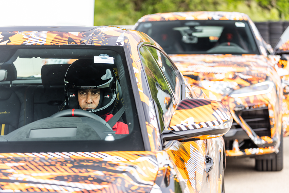 A driver wearing a racing helmet sits inside a camouflaged 2026 Lamborghini Urus Performante prototype, with a second camouflaged prototype visible behind it