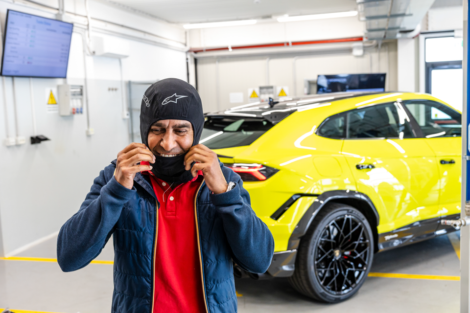 A person putting on an Alpinestars balaclava in a garage, with a yellow Lamborghini Urus Performante SUV visible in the background.