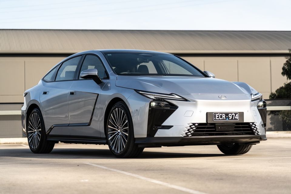 A silver 2026 Lexus ES 350e sedan seen from the front-left side, parked in an open area with a industrial building in the background.