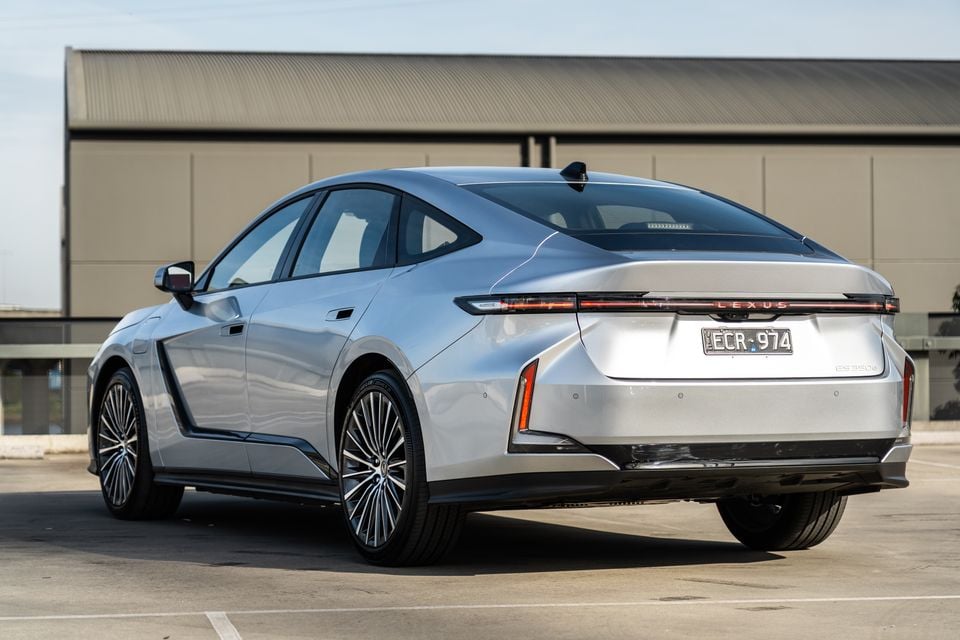 A silver 2026 Lexus ES 350e sedan seen from the rear three-quarter angle in a car park, showing its full-width light bar and Lexus badging