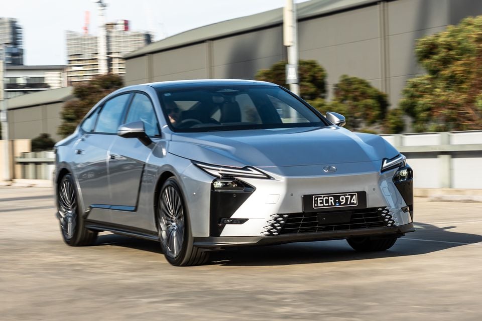 A silver 2026 Lexus ES 350e sedan seen from the front-side angle, driving on an urban road with city buildings in the background.