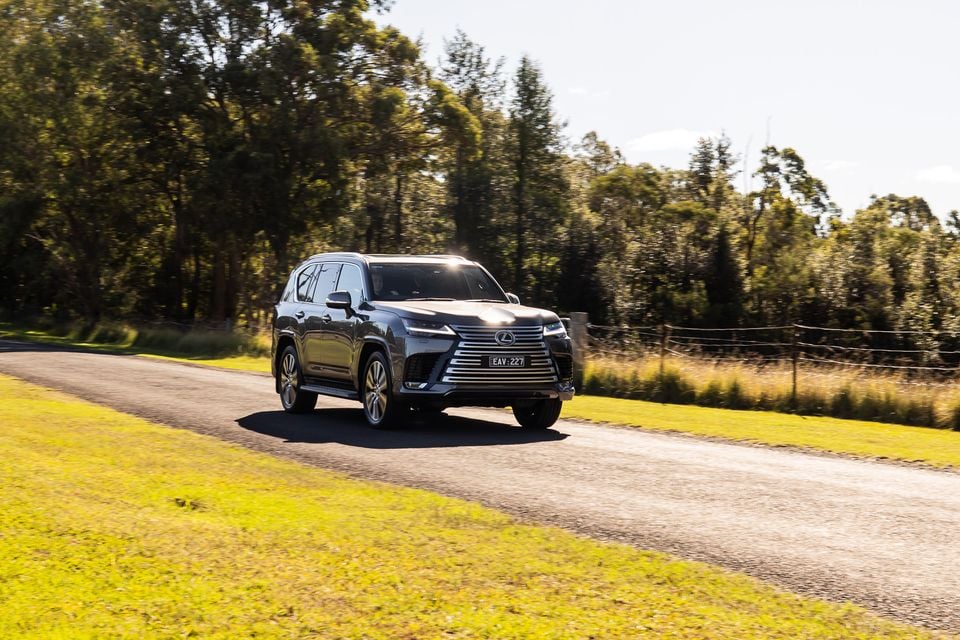 A dark grey 2026 Lexus LX 700h Sports Luxury large SUV driving along a rural road surrounded by trees and green grass