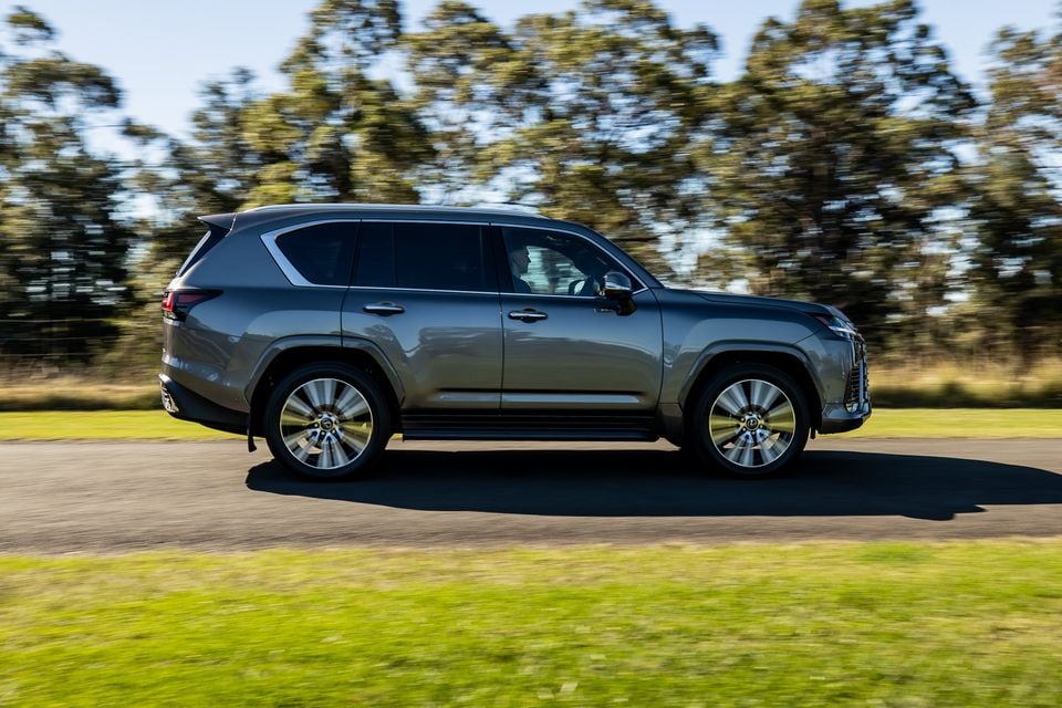 A grey 2026 Lexus LX700h SUV seen from the side while driving on a road surrounded by trees and grass