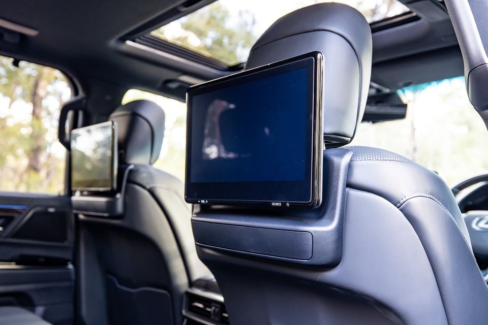 Rear-seat entertainment screens mounted on the front headrests inside a Lexus LX, showing dual screens with black leather seating and a panoramic sunroof visible above
