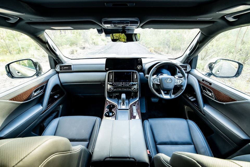 Interior of a Lexus LX700h Sport Luxury showing the dashboard, centre console, leather seats, wood trim, and dual touchscreens, parked on a bush track