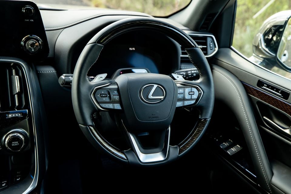 Lexus LX700h leather-trimmed steering wheel with multifunction controls and wood grain trim accents, with the digital instrument cluster visible behind