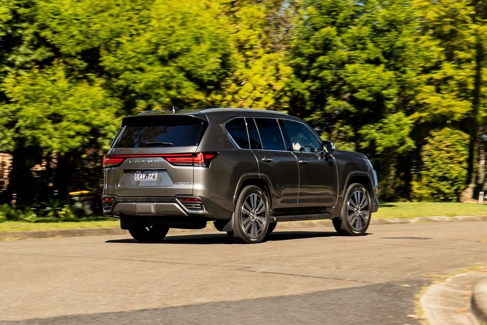 A grey 2026 Lexus LX700h SUV seen from the rear three-quarter angle on a suburban street, surrounded by green trees