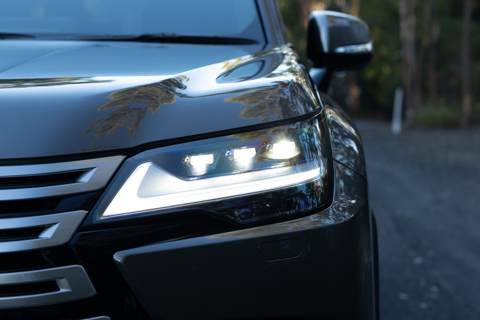 Close-up of the illuminated LED headlight on a dark Lexus LX700h, showing the signature L-shaped daytime running light and spindle grille detail