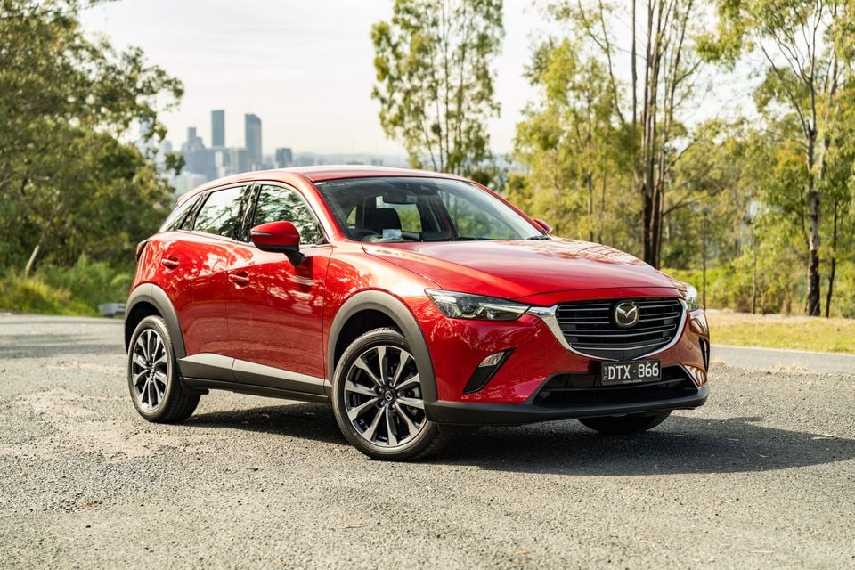 A red Mazda CX-3 small SUV seen from the front-side angle, parked on a gravel road with trees and a city skyline in the background.