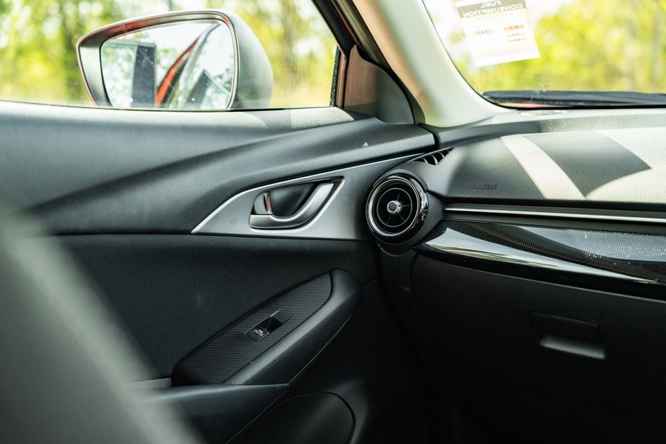 Interior view of a Mazda CX-3 showing the passenger door panel, door handle, side air vent, and dashboard with a fuel consumption sticker visible on the windscreen