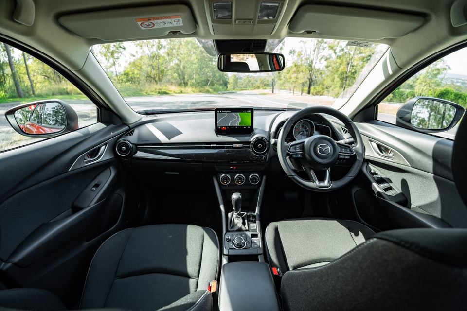 Interior view of a Mazda CX-3 showing the dashboard, centre console, infotainment screen displaying navigation, Mazda steering wheel, and front seats with black fabric upholstery