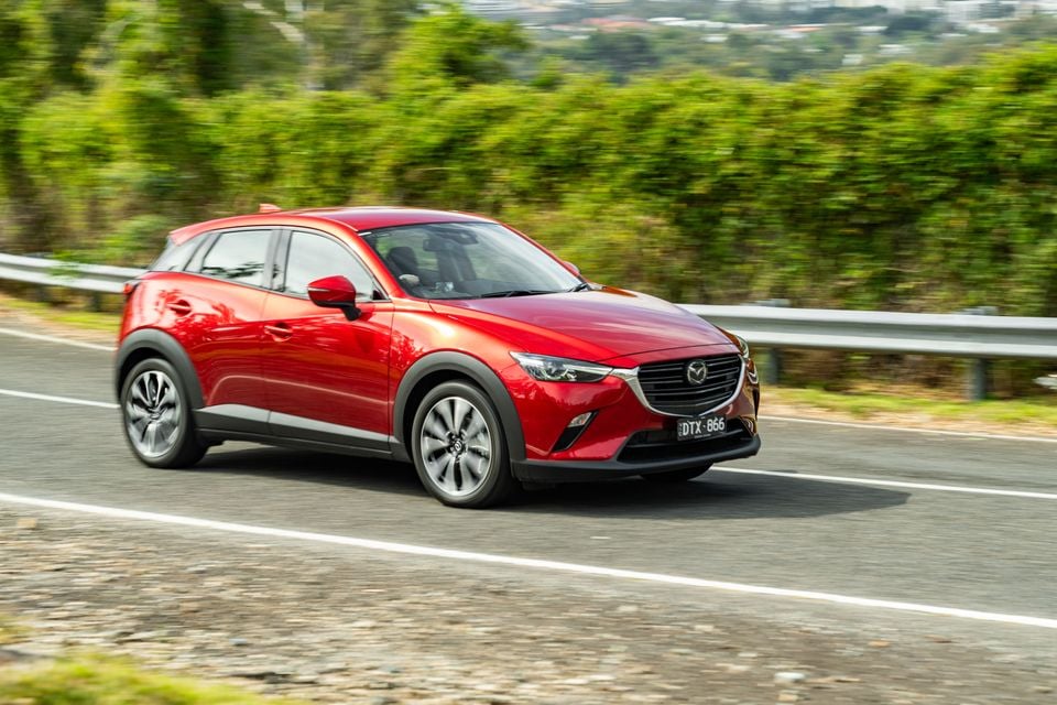 A red Mazda CX-3 small SUV driving on a road surrounded by green trees, seen from the front-side