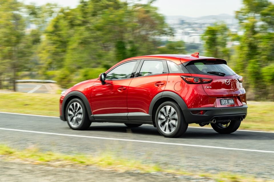 A red Mazda CX-3 small SUV seen from the rear three-quarter angle while driving on a road, with trees and hills in the background