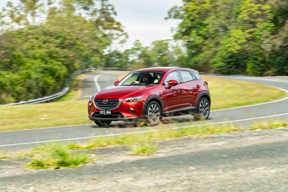 A red Mazda CX-3 SUV being driven along a winding rural road, seen from the front-side