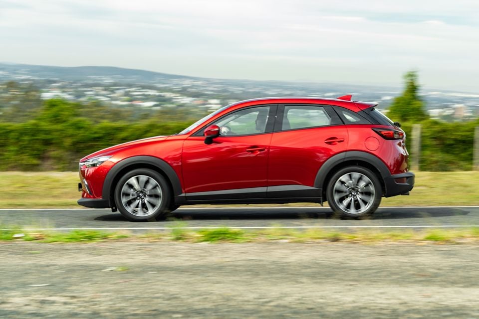 A red Mazda CX-3 small SUV seen from the side while driving along a road with a green hillside and city in the background