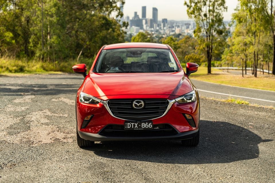 A red 2026 Mazda CX-3 small SUV seen from the front, parked on a road with a city skyline visible in the background.