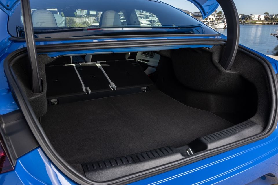 Open boot of a blue 2026 Mercedes-Benz CLA 250 Electric sedan, showing a spacious carpeted cargo area with rear seats partially folded, near a waterfront marina