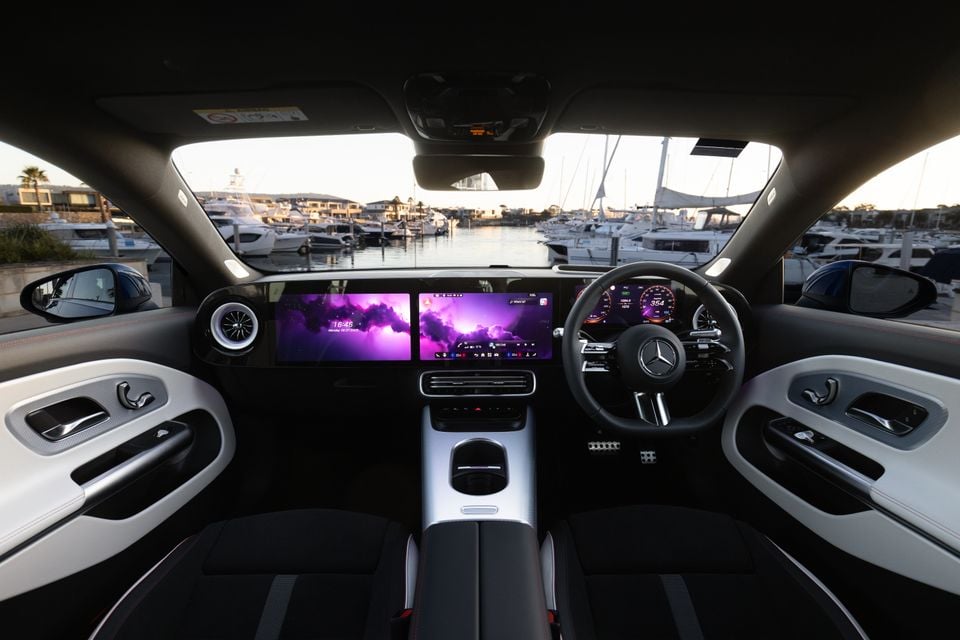 Interior of a 2026 Mercedes-Benz CLA 250 Electric showing the wide dual-screen infotainment display, AMG steering wheel, and dashboard, with a marina visible through the windscreen