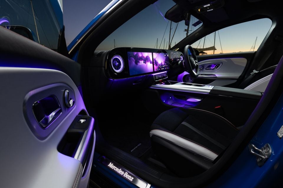 Interior of a 2026 Mercedes-Benz CLA 250 Electric showing the dashboard, wide infotainment screen with purple ambient lighting, AMG-branded seats, and Mercedes-Benz illuminated door sill, photographed at dusk near a marina.