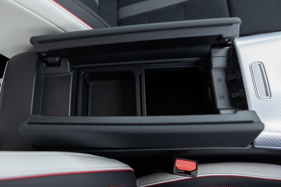 Open centre console storage compartment of a 2026 Mercedes-Benz CLA 250 Electric, showing a wireless charging pad and deep storage bin, with white and black leather seats with red stitching visible