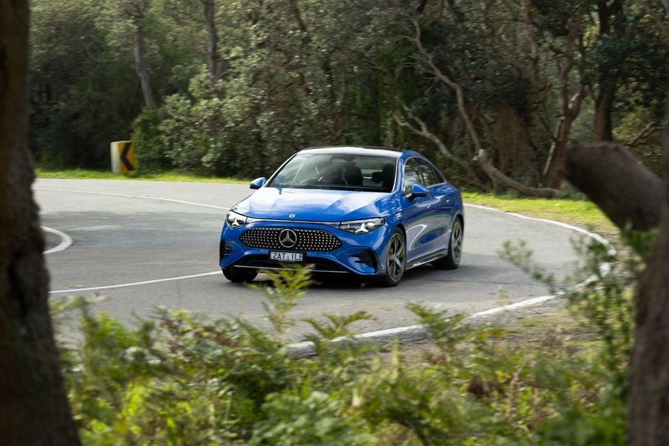 A blue Mercedes-Benz CLA sedan driving around a bend on a tree-lined road, seen from the front