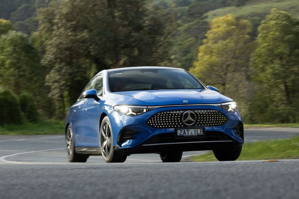 A blue Mercedes-Benz CLA Electric sedan driving on a winding road surrounded by green trees, seen from the front
