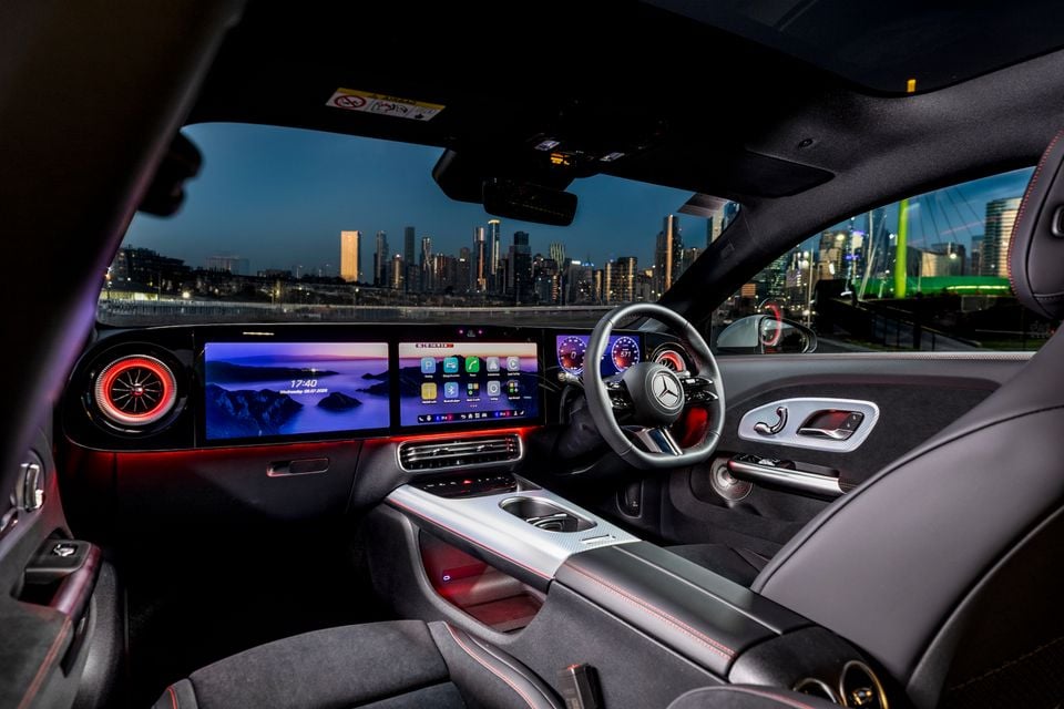 Interior of a 2026 Mercedes-Benz CLA 350 4MATIC Electric showing the wide dual-screen infotainment display, AMG steering wheel, ambient lighting and city skyline at dusk through the windscreen