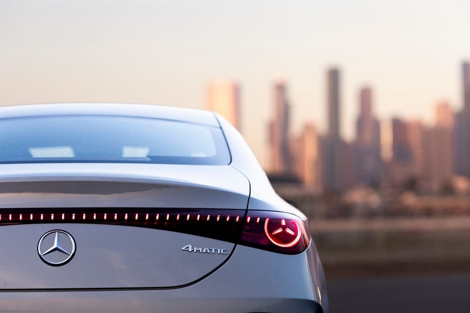 Rear of a silver 2026 Mercedes-Benz CLA 350 4MATIC electric, showing the illuminated tail-lights and badge against a blurred city skyline at dusk.