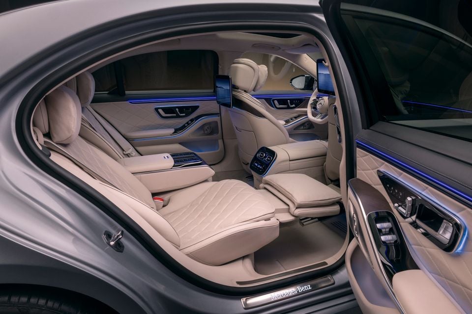 Interior of a Mercedes-Benz S-Class showing luxurious cream quilted leather rear seats with a reclining footrest, rear-seat entertainment screens, ambient blue lighting, and a door panel with controls, viewed through an open rear door.
