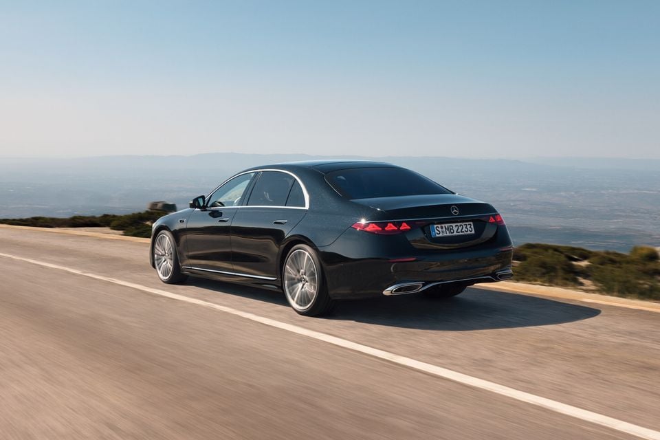 A dark blue 2026 Mercedes-Benz S-Class sedan driving along an open road, seen from the rear three-quarter angle with a hazy valley landscape in the background.