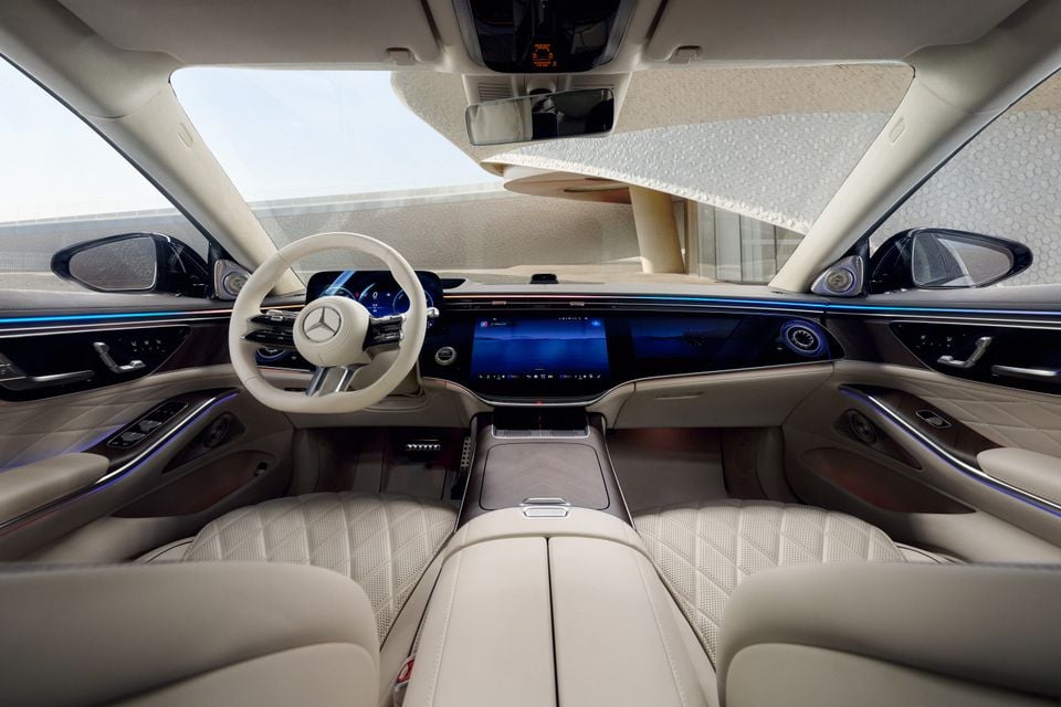 Interior of a 2026 Mercedes-Benz S-Class showing the cream leather seats, large widescreen infotainment display, and circular steering wheel with the Mercedes-Benz star logo