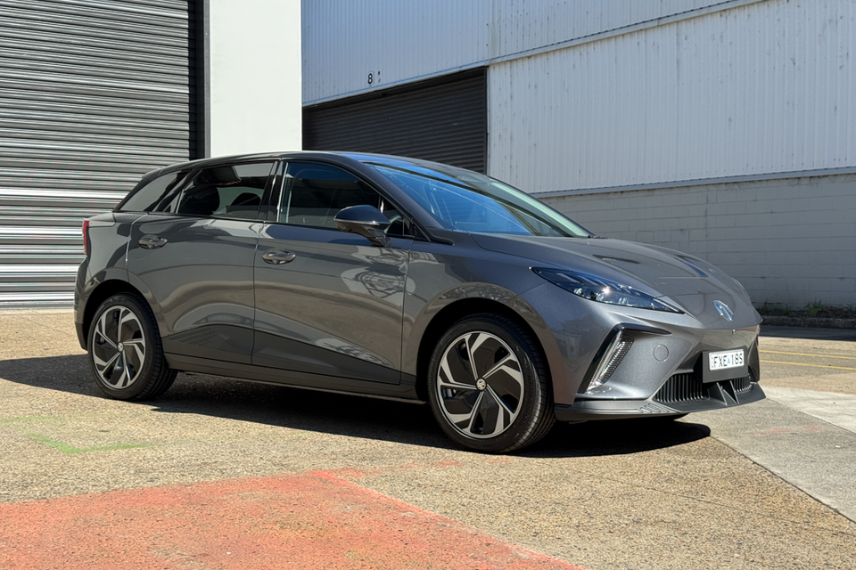 A grey MG4 EV hatchback parked in front of a warehouse roller door, seen from the front-side angle