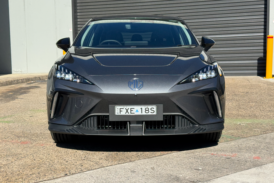 A dark grey MG 4 EV hatchback seen from the front, parked outside a building with a roller door in the background
