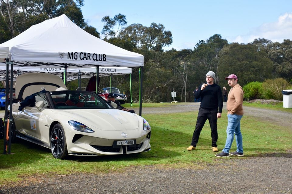 A cream 2026 MG Cyberster convertible parked on grass under an MG Car Club marquee, as two men chat nearby at a car club event
