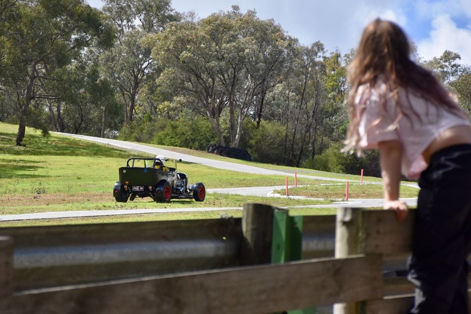 A dark green vintage-style hot rod roadster with red wheels driving away on a grassy hillclimb track as a spectator watches from behind a wooden barrier