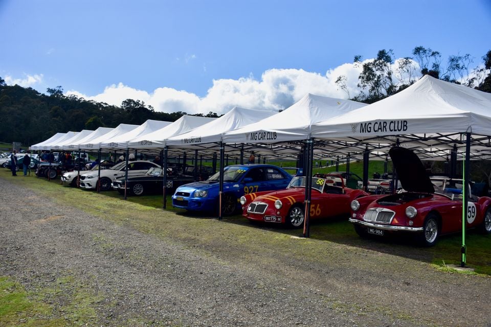 Classic red MGA roadsters, a blue Subaru Impreza race car and other cars parked under MG Car Club marquees at a motorsport event