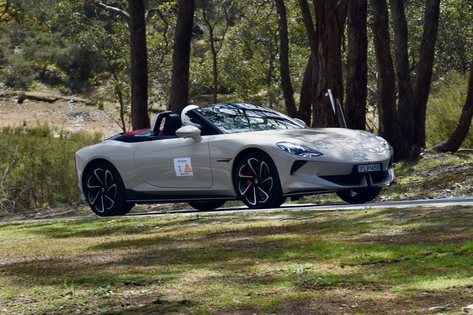 A light grey 2026 MG Cyberster roadster with its roof down being driven along a bush road, seen from the front side