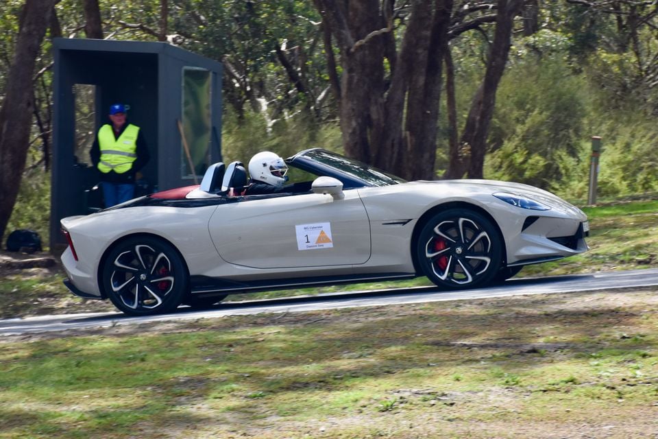 A silver 2026 MG Cyberster convertible driven with the roof down by a helmeted driver at a motorsport event, as a marshal watches