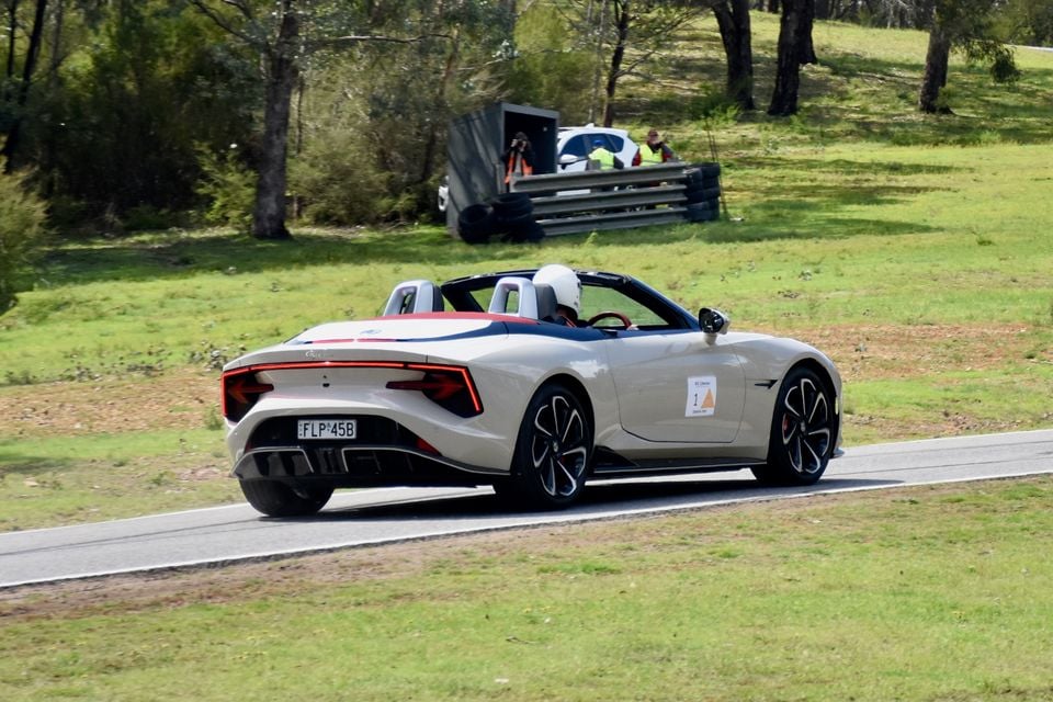 A 2026 MG Cyberster convertible seen from the rear quarter, driven by a helmeted driver on a racetrack with marshals in the background