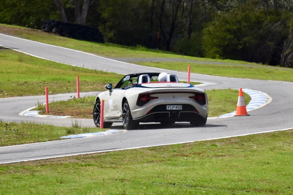A white 2026 MG Cyberster convertible driving through a bend on a race track, seen from the rear with witches hats and markers nearby
