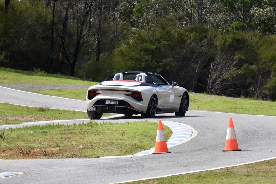 A white 2026 MG Cyberster convertible seen from the rear quarter, driving around a bend on a race track lined with orange traffic cones
