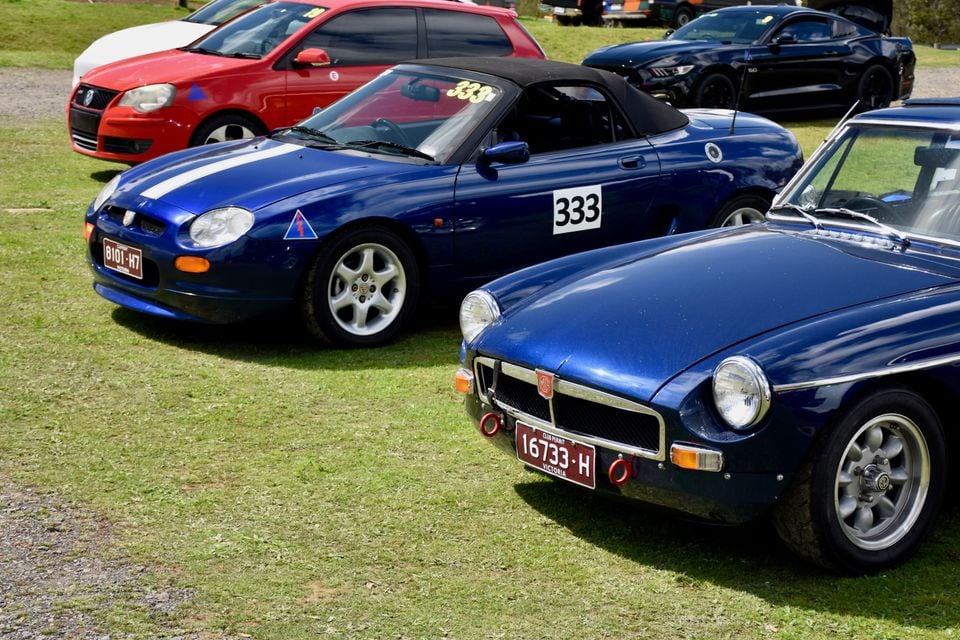 A blue MG F roadster and a classic blue MGB parked side by side on grass at a car club event, with other cars behind