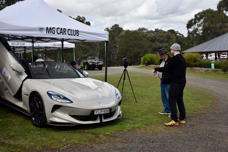 A pearl white 2026 MG Cyberster with its scissor door raised, parked under an MG Car Club marquee as two men look on beside a camera tripod
