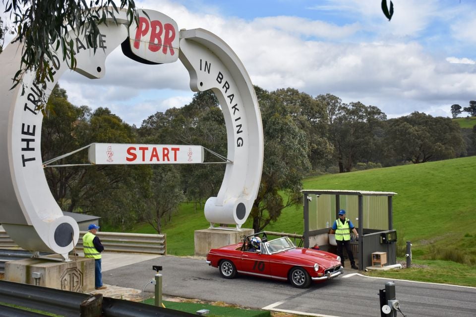 A classic red MGB roadster with racing number 10 waiting at the start line beneath a PBR arch at a hillclimb, with marshals in hi-vis vests