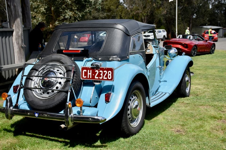 A light blue vintage MG roadster with a black soft top and rear-mounted spare wheel parked on grass, seen from the rear, with a red MG Cyberster behind