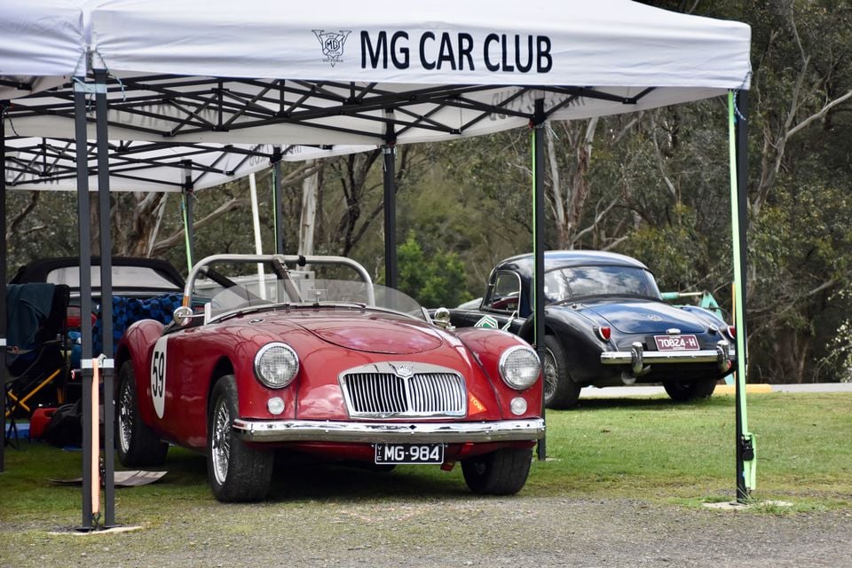 A red classic MG MGA roadster with race number 59 parked under an MG Car Club marquee, with a black MGA coupe parked behind on the grass