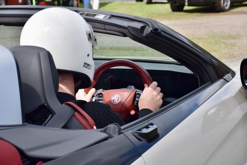 Helmeted driver gripping the red steering wheel of a white 2026 MG Cyberster convertible, seen from behind over the shoulder with the roof down